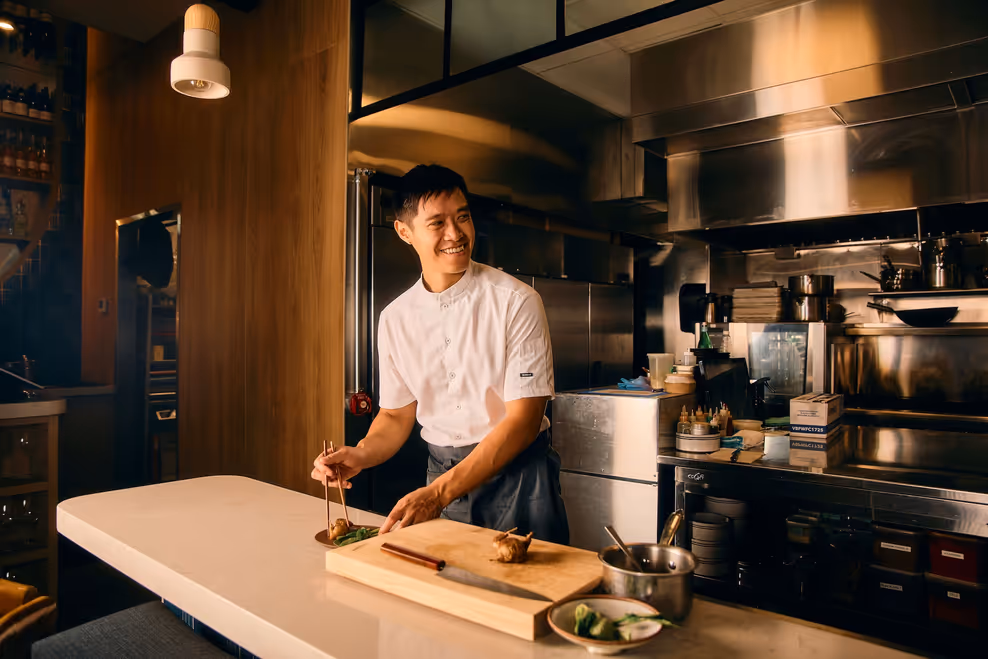 Chef Steven Che smiling as he plates a dish at the counter of Haisan's open kitchen